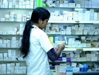 Trusted Indian pharmacist standing in a pharmacy with medicine shelves.